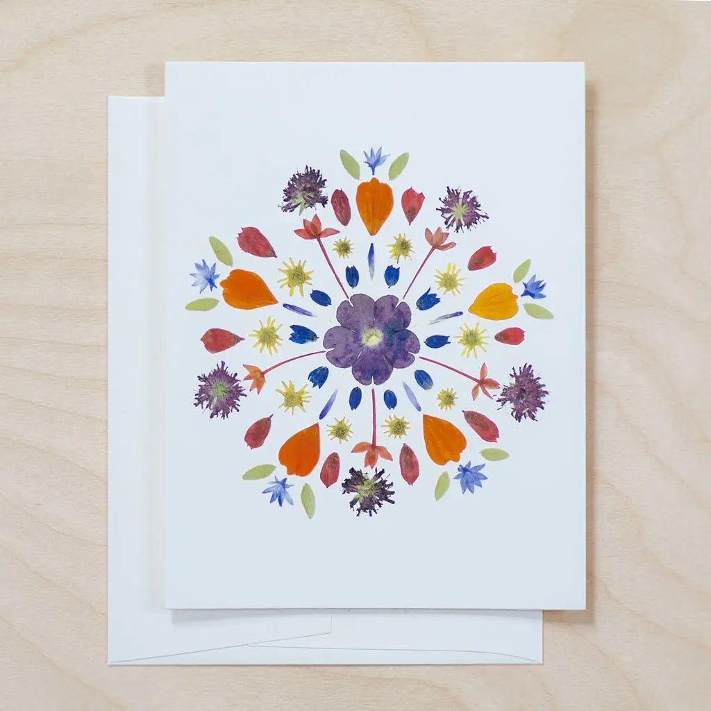 The PETAL PEOPLE PRESS - MANDALA CARD by PETAL PEOPLE PRESS features a vibrant pressed flower and leaf mandala, styled with scissors, a bowl of paperclips, and green leaves on a white background.