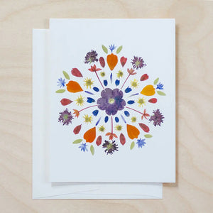 The PETAL PEOPLE PRESS - MANDALA CARD by PETAL PEOPLE PRESS features a vibrant pressed flower and leaf mandala, styled with scissors, a bowl of paperclips, and green leaves on a white background.