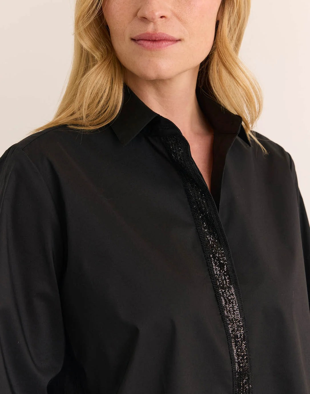 A woman wearing a black HINSON WU - ESTHER LONG SLEEVE SHIRT WITH SEQUIN TRIM under a black velvet blazer, paired with olive green trousers, black high-heeled sandals, and holding a black clutch.