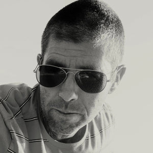 The CADDIS - MABUHAY SUNGLASSES by CADDIS feature a thin gold wire frame, scratch-resistant dark lenses, and brown temple tips, displayed against a white background.