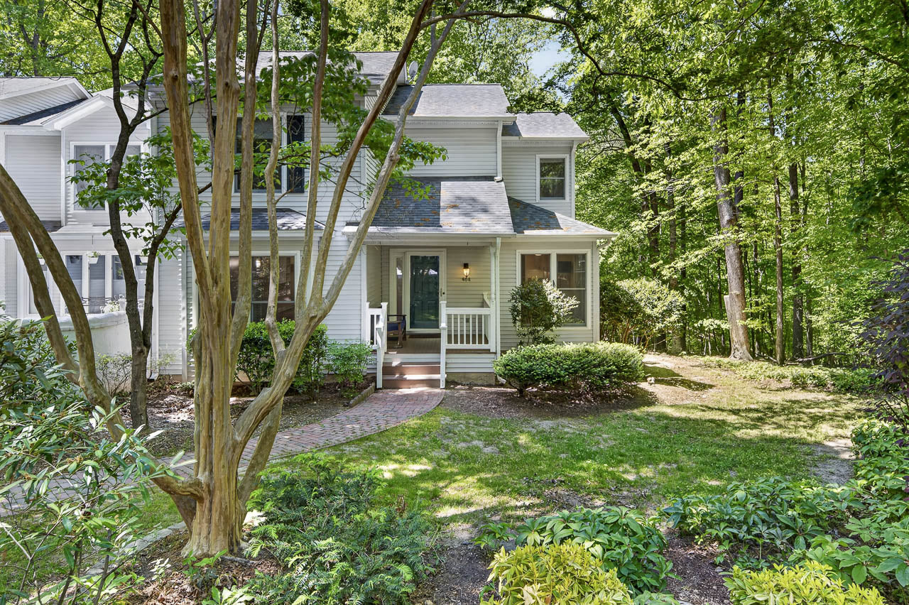 A two-story light gray house with white trim, a front porch, and a brick walkway surrounded by green trees, shrubs, and landscaped garden in a wooded setting.