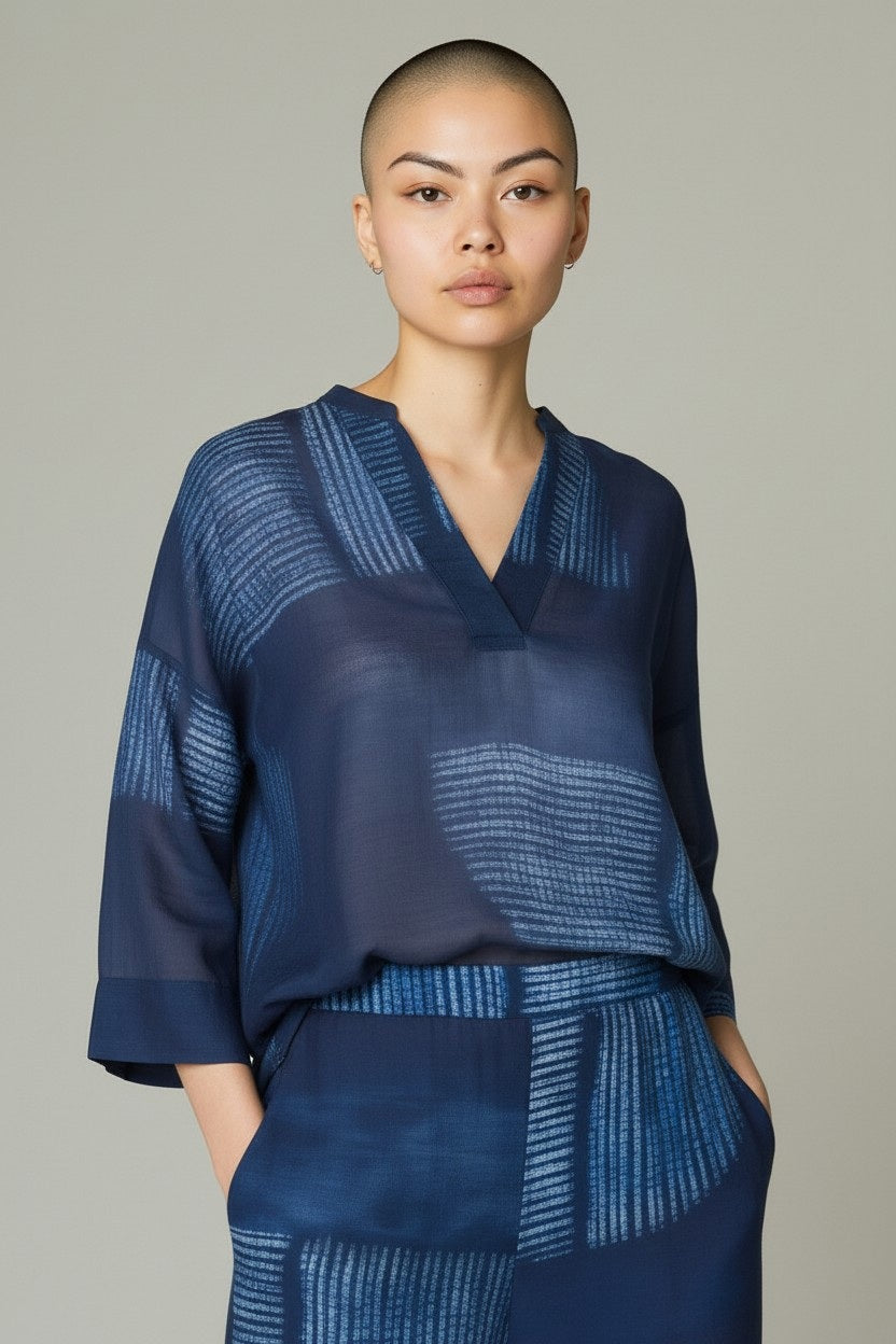 A person with a shaved head models the SANDRO FERRONE SANRO FERRONE - PRINTED BLOUSE and matching pants, featuring light blue patterned stripes. They stand with hands in pockets against a plain light gray background, showcasing a relaxed look.