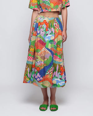 A woman stands against a plain background wearing green sandals and the MIRTO 1956 MULTICOLOR PRINT BALLOON SKIRT, featuring vibrant floral and peacock patterns, paired with a matching colorful crop top and straw hat.