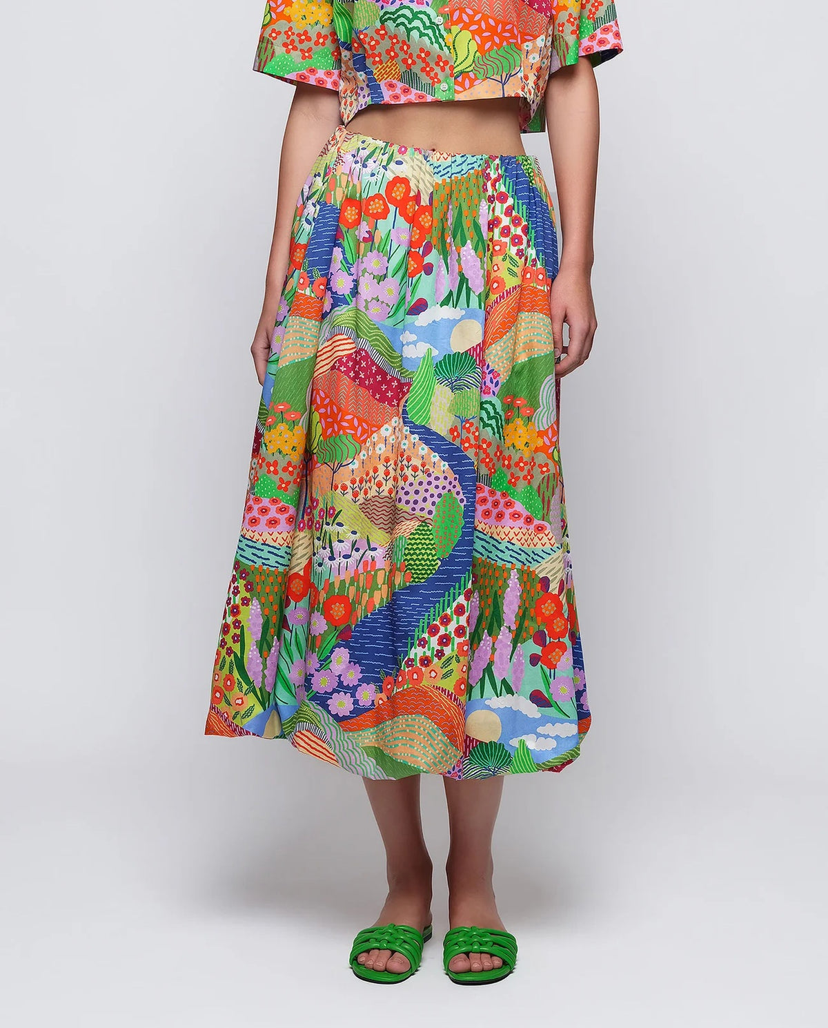 A woman stands against a plain background wearing green sandals and the MIRTO 1956 MULTICOLOR PRINT BALLOON SKIRT, featuring vibrant floral and peacock patterns, paired with a matching colorful crop top and straw hat.