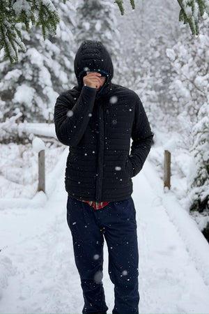 Wearing the RELWEN - WINDZIP HOOD IN BLACK FADE, a black water-resistant jacket with recycled polyester insulation from RELWEN, a person stands on a snowy path in blue pants, covering their face with their hand. Snow falls gently as snow-laden trees fill the background.