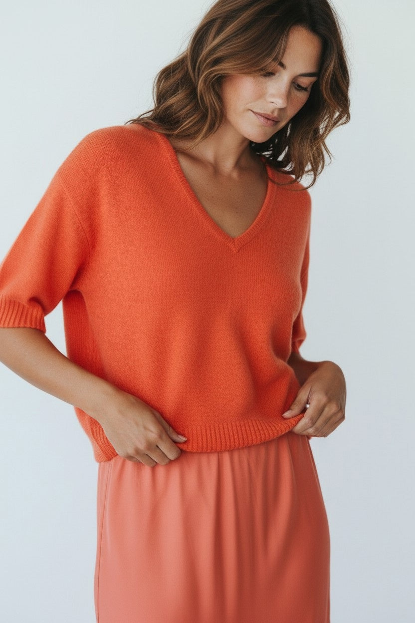 A woman with wavy brown hair wears the ROSE & CROWN Cashmere V Neck Short Sleeve Sweater in orange, paired with a coral skirt, looking down with a relaxed expression against a light background.