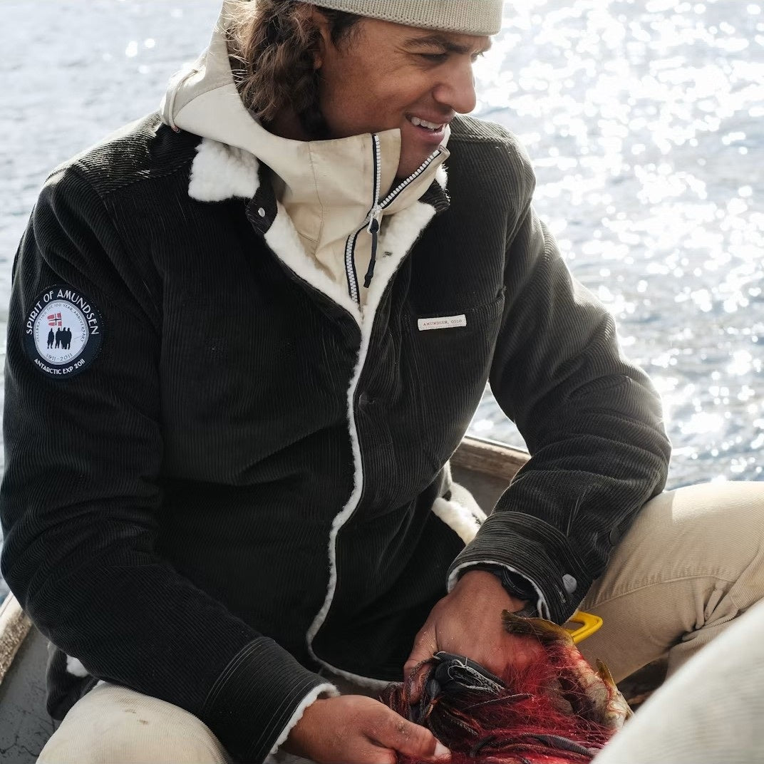 A person in a warm jacket and beanie smiles while holding a red-feathered bird, possibly on a boat, wearing the AMUNDSEN SPORTS - HARVESTER OVERSHIRT.