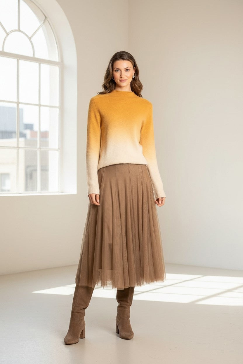 A woman stands in a sunlit room wearing a yellow-to-cream ombre sweater, the JESSIE LIU Tulle Midi Skirt with Merino Wool Lining in Brown, and brown knee-high boots. She smiles softly as natural light streams through a large window, filling the space.