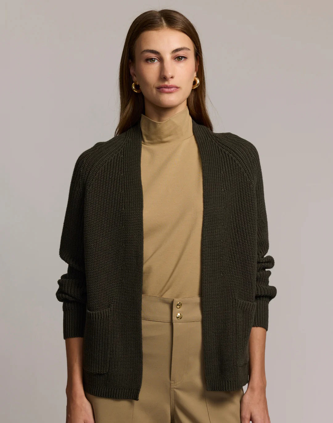 A woman with straight brown hair, in a tan turtleneck and high-waisted tan pants with gold buttons, wears the HINSON WU ANGIE Long Sleeve Cardigan in dark olive green, standing arms crossed against a plain light gray background.
