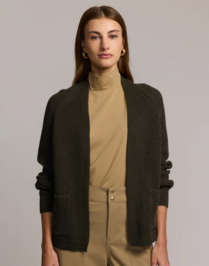 A woman with straight brown hair, in a tan turtleneck and high-waisted tan pants with gold buttons, wears the HINSON WU ANGIE Long Sleeve Cardigan in dark olive green, standing arms crossed against a plain light gray background.