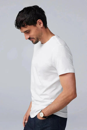 A man with short dark hair and a beard wears the Mack Weldon Silver Crew Neck T-Shirt in Desert Taupe, paired with dark jeans and a wristwatch. He stands against a light gray background, looking slightly to the side with a neutral expression.