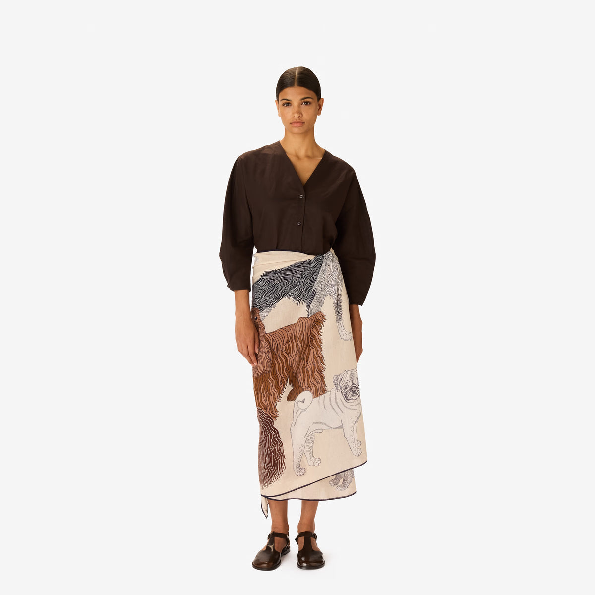 A woman in a dark outfit stands with her hands in her pockets, draped in the INOUI EDITIONS - TOUTOU Scarf in White by INOUI, featuring intricate dark brown and navy animal illustrations. She is set against a plain white background.
