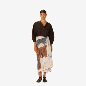 A woman in a dark outfit stands with her hands in her pockets, draped in the INOUI EDITIONS - TOUTOU Scarf in White by INOUI, featuring intricate dark brown and navy animal illustrations. She is set against a plain white background.