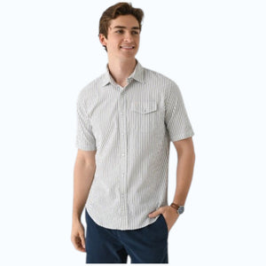 A young man, smiling with one hand in his pocket, wears CRIQUET’s Short Sleeve Performance Seersucker Pearl Snap Shirt in Sage and navy blue pants.
