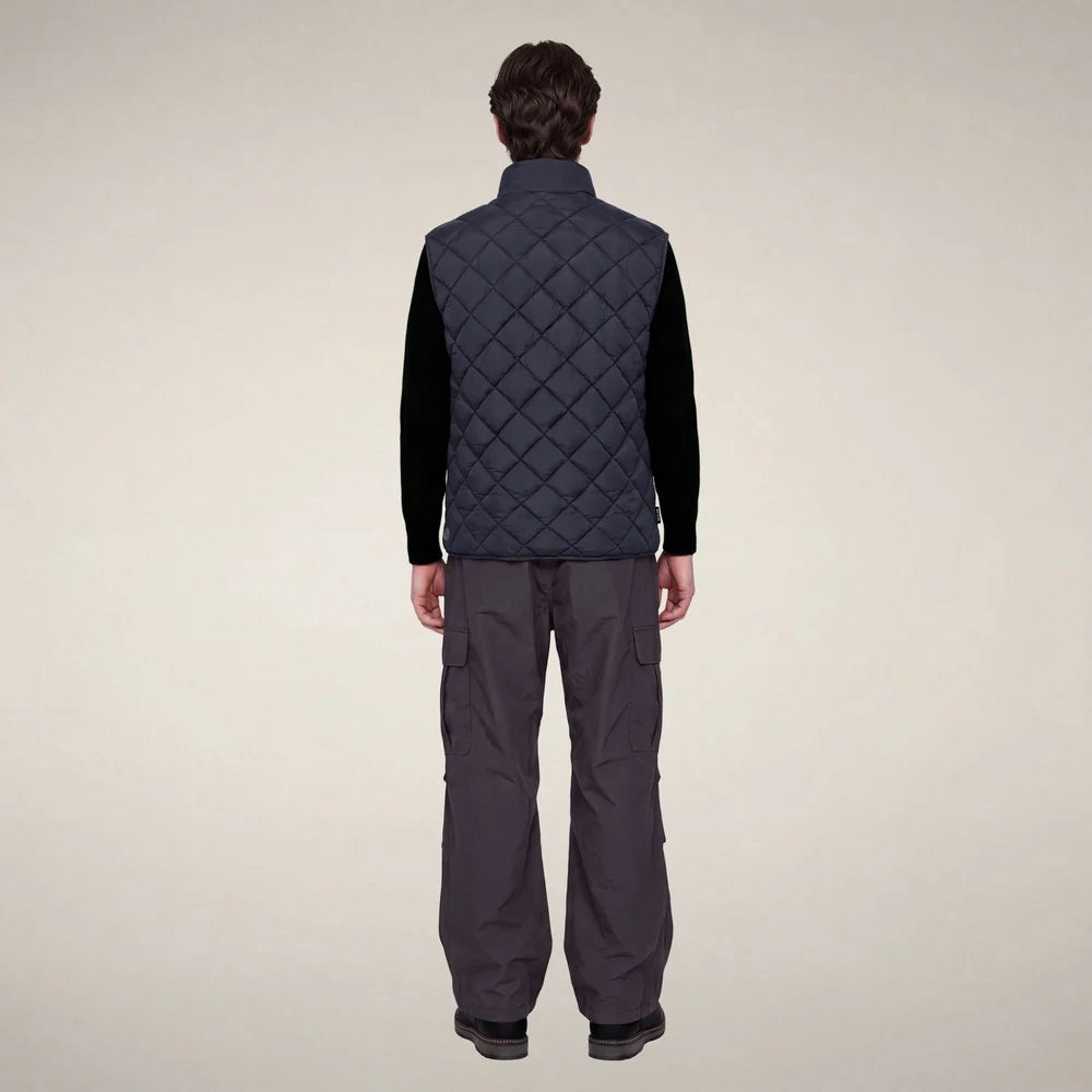A person stands facing forward, wearing the SAVE THE DUCK - FABRICE Puffer Vest in Blue Black from SAVE THE DUCK, featuring recycled Plumtech insulation, layered over a black long-sleeve shirt and gray pants. They have a neutral facial expression and are posed against a plain white background.