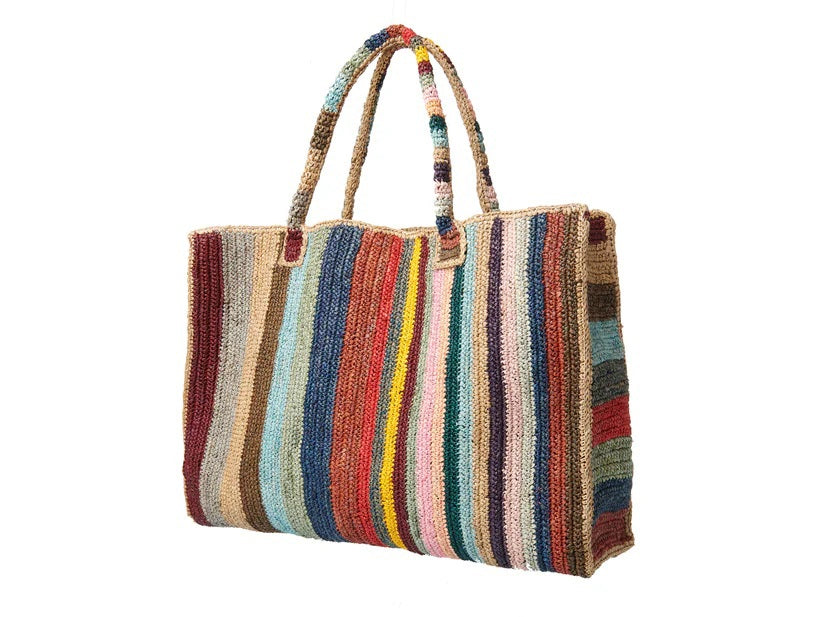 A person in blue jeans and a white shirt holds the GABRIELE FRANTZEN - RAFFIA WEEKENDER BAG CIRCUS, a large, hand-crocheted tote with two handles featuring vibrant vertical stripes and showcasing natural fiber craftsmanship.