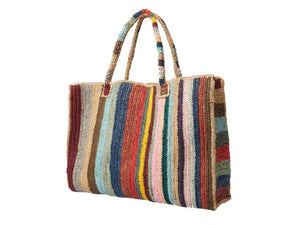 A person in blue jeans and a white shirt holds the GABRIELE FRANTZEN - RAFFIA WEEKENDER BAG CIRCUS, a large, hand-crocheted tote with two handles featuring vibrant vertical stripes and showcasing natural fiber craftsmanship.