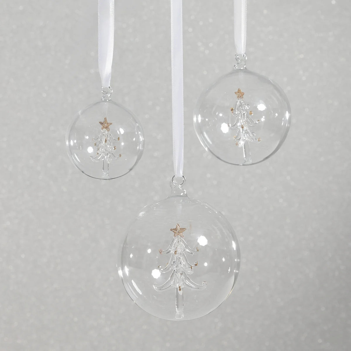 The ZODAX CLEAR GLASS BALL ORNAMENT WITH TREE INSIDE is a 4-inch ornament featuring a white glass Christmas tree topped with a gold star, suspended by a beige ribbon, beautifully displayed against a white background.