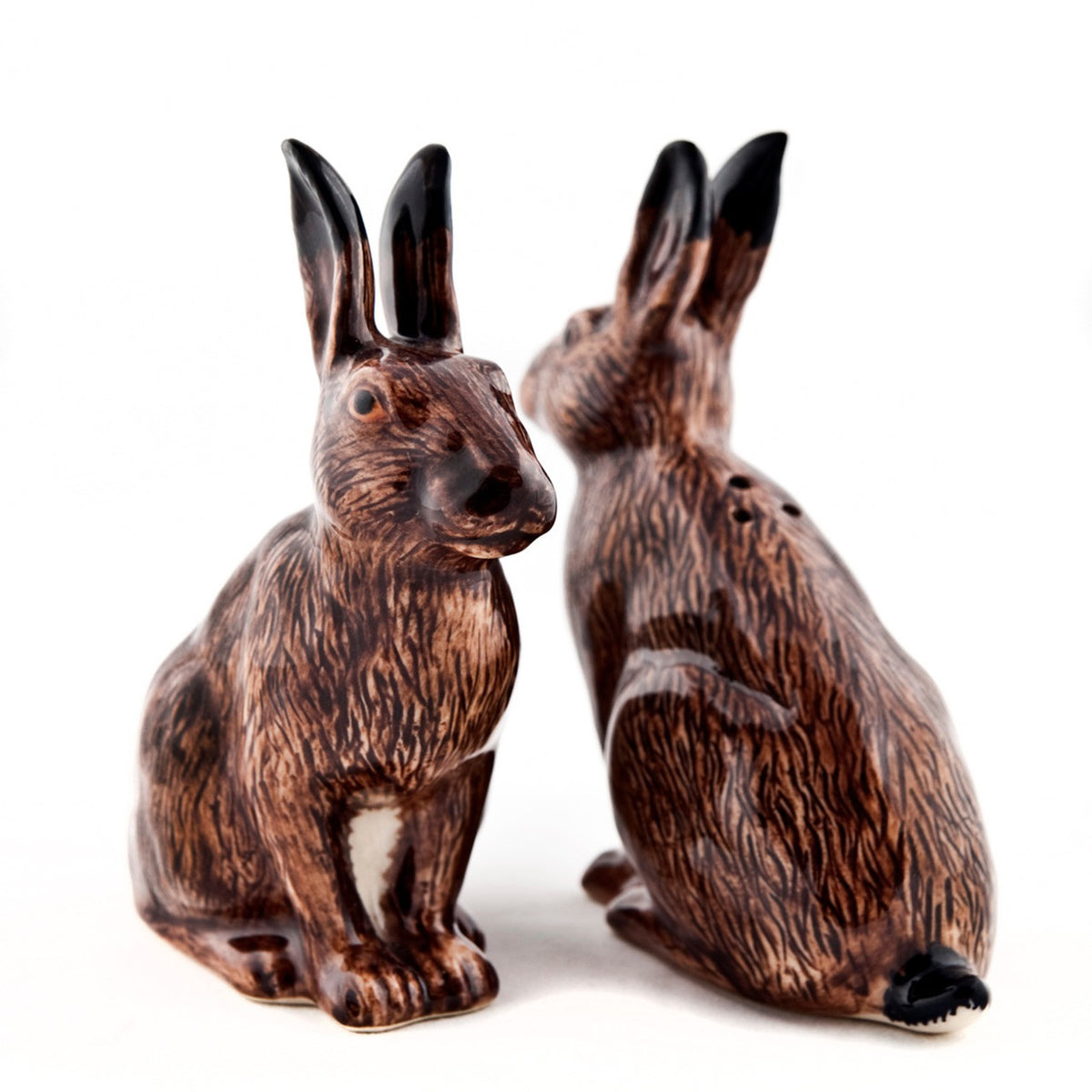 The QUAIL CERAMICS - HARE SALT AND PEPPER SET by QUAIL DESIGNS features two handpainted ceramic rabbit shakers with black-tipped ears—one facing forward, the other turned slightly to the side—on a white background.