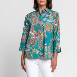 The individual is wearing the Hinson Wu Xena Shirt, a teal paisley print blouse with 3/4 sleeves and a collar. The blouse showcases an intricate design featuring vibrant colors like orange, purple, and blue. They have paired it with white pants against a plain white background.