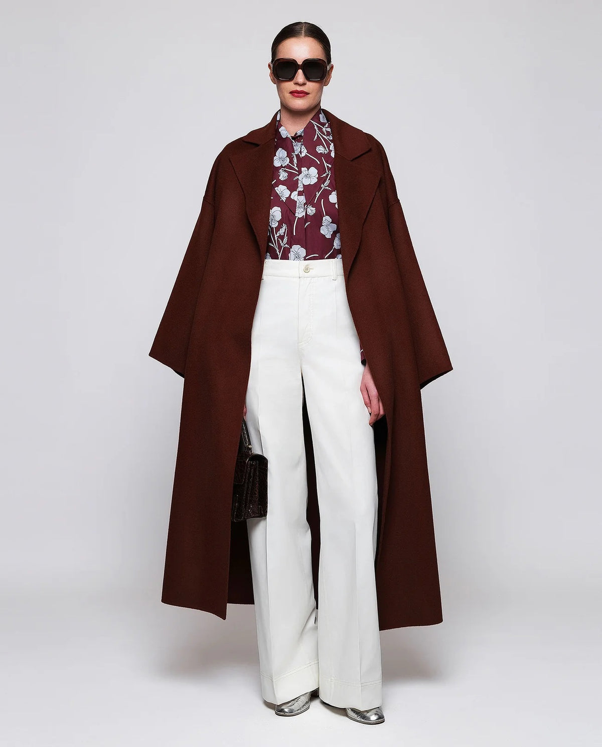 A woman stands by a window in a well-lit modern room, wearing the MIRTO 1956 Floral Print Blouse with Tie Neck in burgundy and high-waisted white pants, her hands casually tucked into her pockets.
