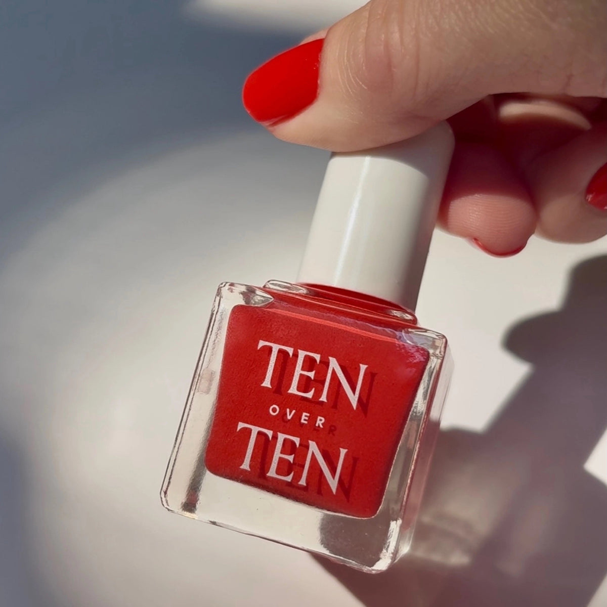 A glass bottle of TENOVERTEN - NAIL POLISH from the brand TEN OVER TEN, featuring a white cap and a dusty pink nail color. The front label displays "TEN OVER TEN" in white uppercase letters. This vegan, eight-free formula ensures both beauty and peace of mind. The background is plain and light-colored.