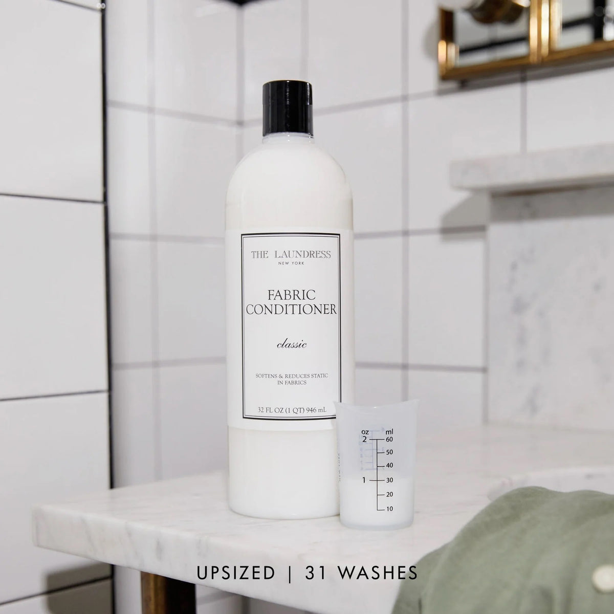A white bottle of THE LAUNDRESS - FABRIC CONDITIONER 32OZ with a &quot;Classic&quot; scent label. This 32 fluid ounce (946 milliliters) fabric conditioner by THE LAUNDRESS promises to soften fabrics and reduce static. The bottle features a black cap, set against a light gray background.