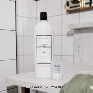 A white bottle of THE LAUNDRESS - FABRIC CONDITIONER 32OZ with a "Classic" scent label. This 32 fluid ounce (946 milliliters) fabric conditioner by THE LAUNDRESS promises to soften fabrics and reduce static. The bottle features a black cap, set against a light gray background.