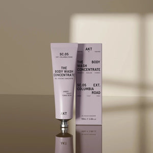 A tube of AKT LONDON BODY WASH CONCENTRATE COLUMBIA ROAD—a luxurious concentrated body wash enriched with olive squalane—stands upright in front of its matching box on a neutral surface, with a soft shadow in the background.