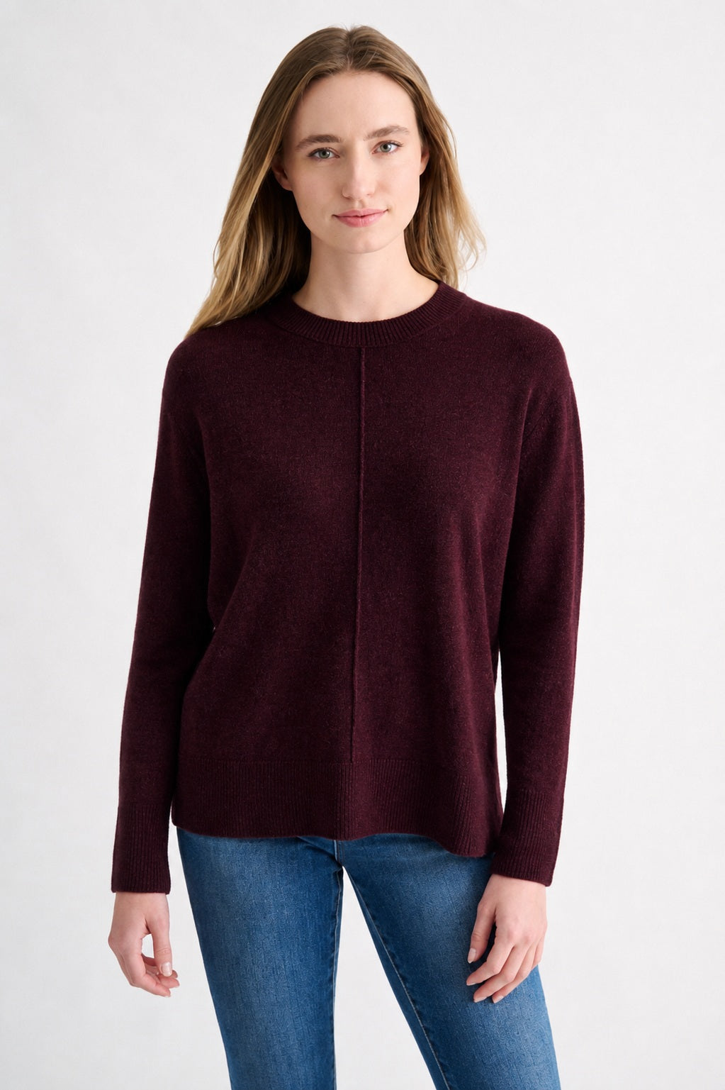 A person with long light brown hair wears the KINROSS CASHMERE Hi Low Seamed Crew Neck Cashmere Sweater in Cordovan—featuring a relaxed fit—paired with blue jeans, standing against a plain light background and facing forward with a neutral expression.
