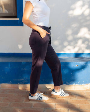 A person wears the MERSEA - FRENCHIE CIGARETTE PANT by MERSEA, paired with a white top and gray sneakers, standing on sandy beach ground with ocean waves and blue sky behind them, hands in their pockets.