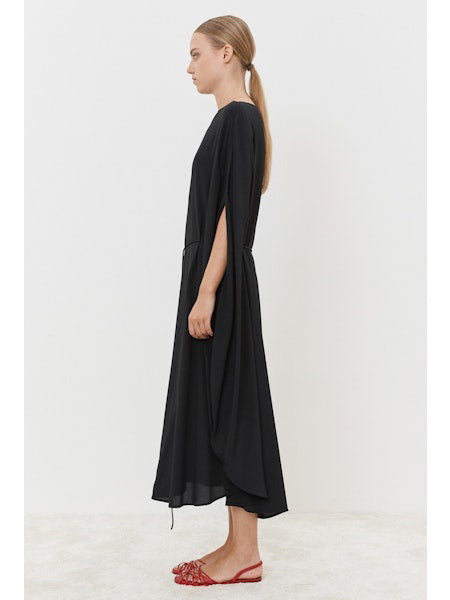 A person stands gracefully on a light-colored floor, wearing the MEIMEIJ Silk Blend Dress with cape-like sleeves, showcasing a modern classic look. The outfit is paired with red, open-toed flat shoes against a simple white backdrop.