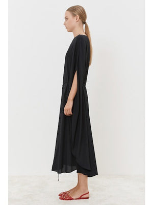 A person stands gracefully on a light-colored floor, wearing the MEIMEIJ Silk Blend Dress with cape-like sleeves, showcasing a modern classic look. The outfit is paired with red, open-toed flat shoes against a simple white backdrop.