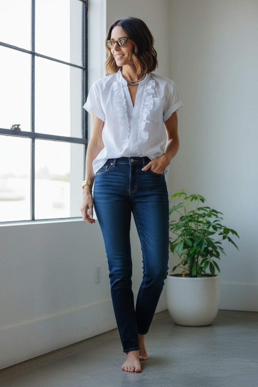 A woman wears the KMJ KIKI SHIRT, a black stand collar blouse with front ruffles, tucked into high-waisted blue jeans. She accessorizes with layered gold jewelry and red lipstick, hands resting on her hips. Brand: KMJ.