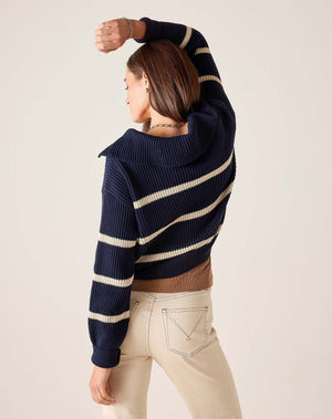 Wearing the MERSEA Camden Sailor Sweater in Navy Cream—adorned with classic white stripes—a woman stands in a sunlit room with expansive windows, smiling as she pairs the look with cream high-waisted pants, effortlessly channeling coastal Maine charm.