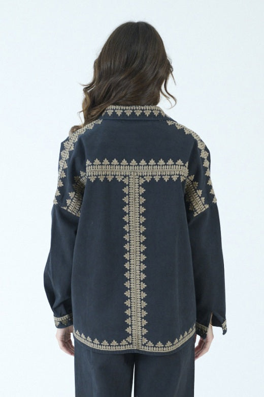 A woman with long brown hair stands indoors in front of large windows, embodying effortless style in the STORIATIPIC ROXY JACKET IN NOIR by STORIATIPIC, featuring gold embroidery on 100% cotton. She pairs the jacket with a patterned shirt, dark pants, and black loafers, mindful to follow proper cotton care such as gentle machine wash.