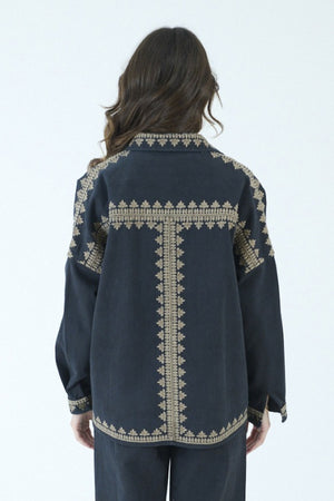 A woman with long brown hair stands indoors in front of large windows, embodying effortless style in the STORIATIPIC ROXY JACKET IN NOIR by STORIATIPIC, featuring gold embroidery on 100% cotton. She pairs the jacket with a patterned shirt, dark pants, and black loafers, mindful to follow proper cotton care such as gentle machine wash.