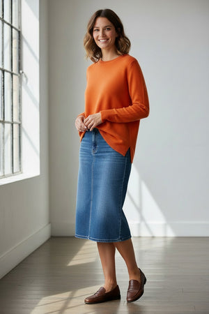 A woman with wavy dark hair wears the BASE MILANO - BOXY OVERSIZED CREWNECK SWEATER in mustard yellow and a blue denim skirt, standing against a plain light background with one arm crossed and a neutral expression.