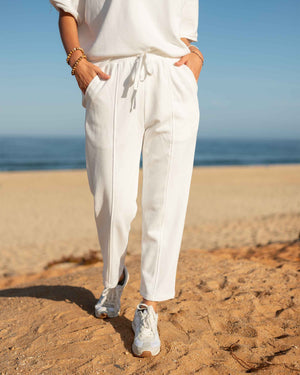 A person wears the MERSEA - FRENCHIE CIGARETTE PANT by MERSEA, paired with a white top and gray sneakers, standing on sandy beach ground with ocean waves and blue sky behind them, hands in their pockets.