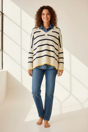 Wearing MERSEA’s Montauk Colorblock V Neck Sweater in Cream Stripe, a person with sunglasses and a scarf stands on a boat, holding the railing against a backdrop of calm blue water and clear sky.