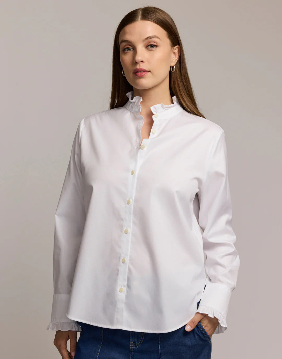 A woman with long brown hair wears the HINSON WU Quinn Long Sleeve No Iron Cotton Shirt, featuring a ruffled collar and cuffs, paired with blue jeans. She stands against a light background—showcasing effortless everyday style.