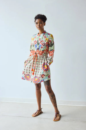 A woman with short curly hair and tattoos on her left leg stands indoors by a plain wall, wearing the HOPE FOR FLOWERS - BAND COLLAR TUNIC IN CUSTARD FLORAL from HOPE FOR FLOWERS, paired with sandals.