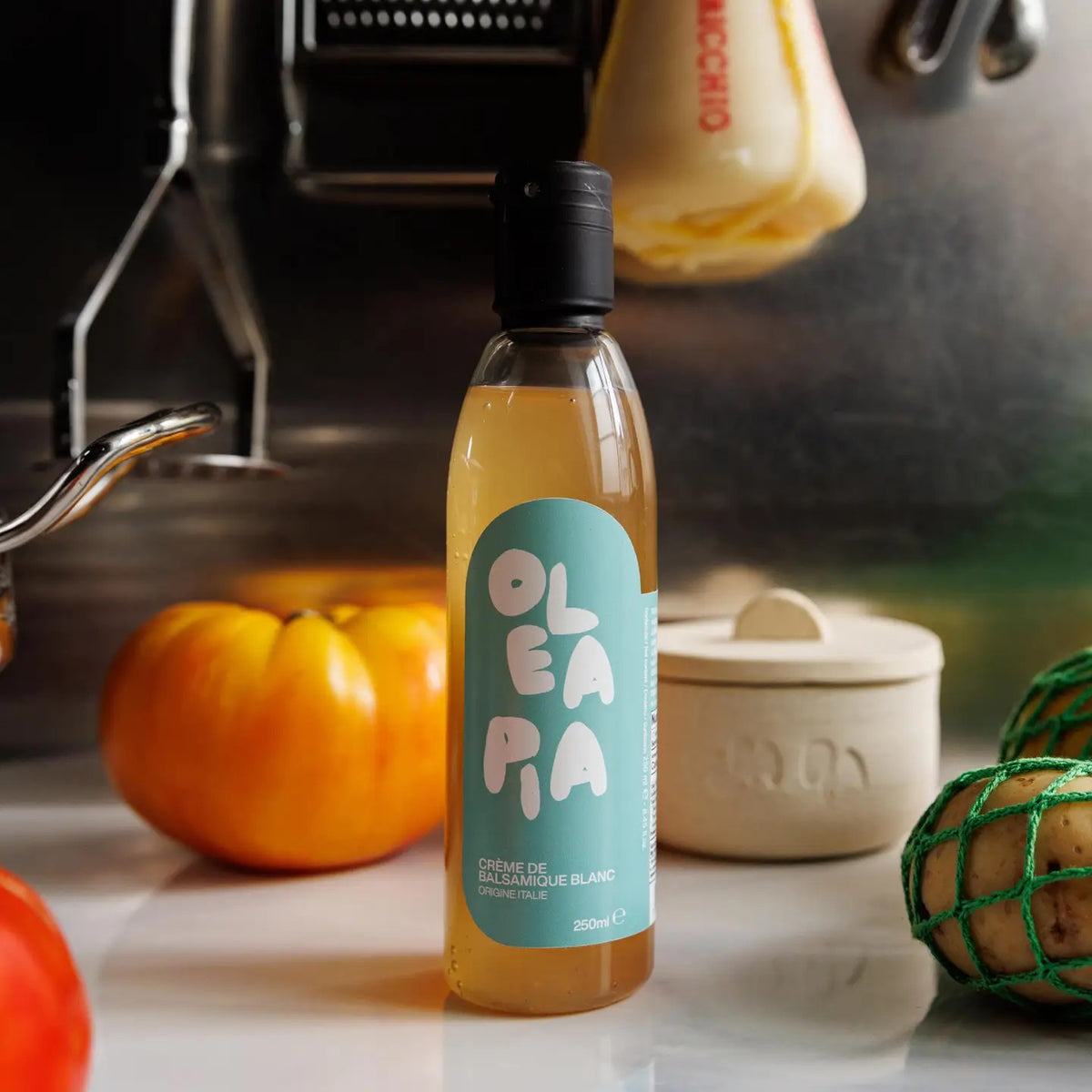 A bottle of OLEA PIA - WHITE BALSAMIC VINEGAR CREAM sits elegantly on a kitchen counter, enhancing the culinary ambiance with its sophistication. It is surrounded by an orange tomato, a green-wrapped item, a pot, and various kitchen utensils in the background. The bottle's label showcases a light blue design with white text.