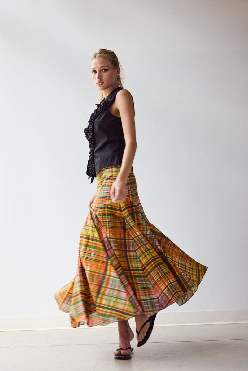 A woman stands indoors against a plain wall, wearing the HOPE FOR FLOWERS Circle Skirt in Spring Plaid, crafted from GOTS certified organic cotton lawn. She pairs it with a sleeveless black button-up top, black sandals, and has her hair tied back.