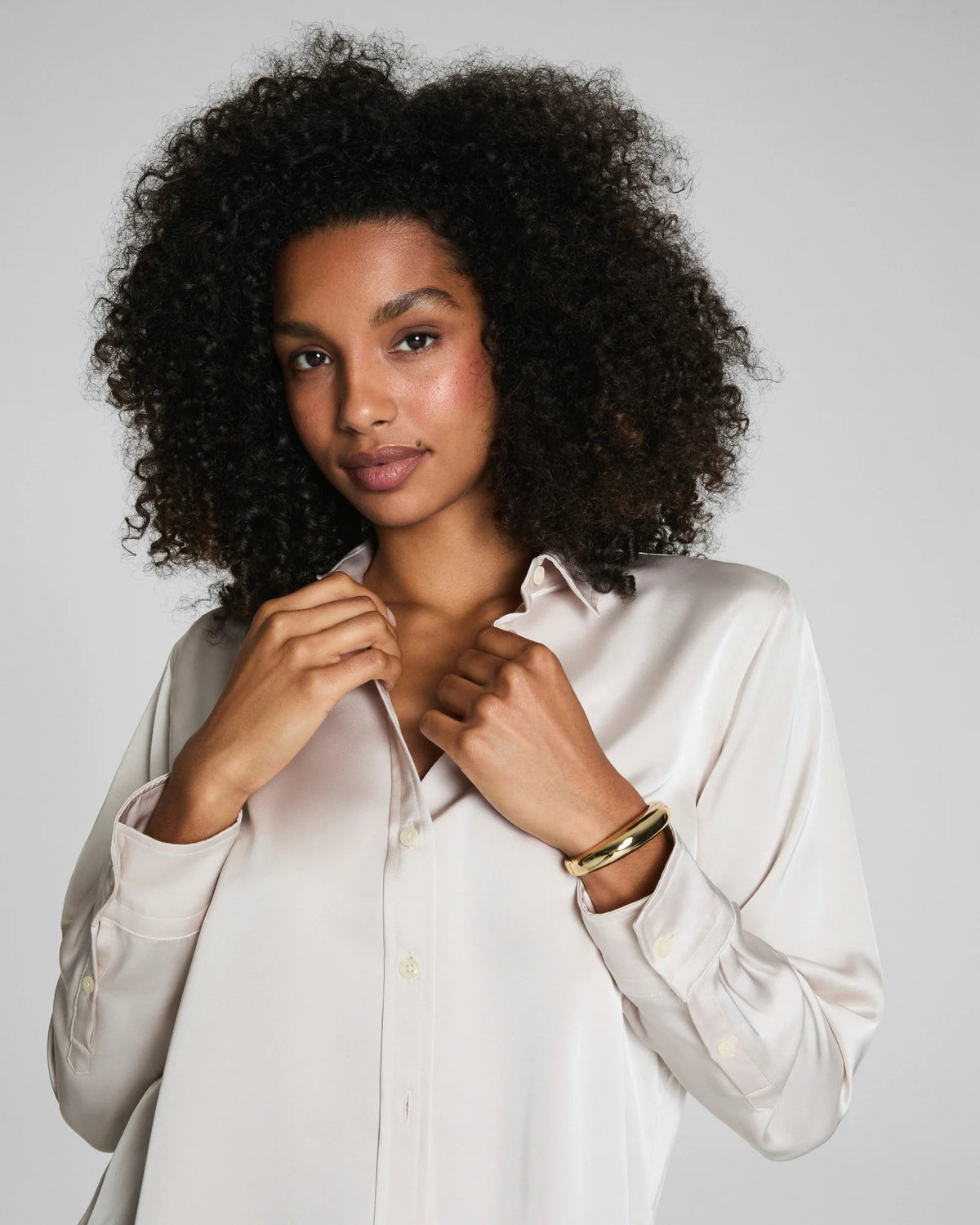 A woman with curly hair wears the SPANX Silky Smooth Button Down Shirt in a relaxed tailored fit and black pants, turning slightly as she looks over her shoulder against a plain gray background.