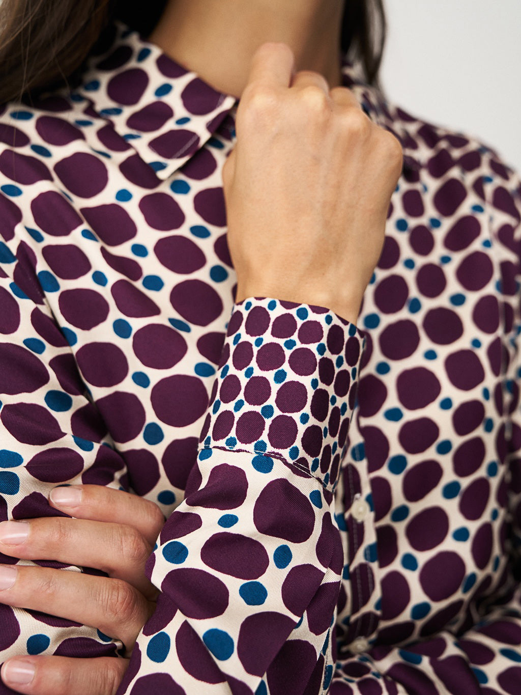 A person with straight brown hair adjusts the collar of the 0039 ITALY SIA BLOUSE, which features a geometric circle print with large green spots and small burgundy dots on cream. The side view highlights the blouse&#39;s viscose sleeve.
