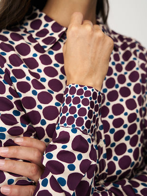 A person with straight brown hair adjusts the collar of the 0039 ITALY SIA BLOUSE, which features a geometric circle print with large green spots and small burgundy dots on cream. The side view highlights the blouse's viscose sleeve.