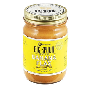 A glass jar of BIG SPOON ROASTERS - WAG BUTTER BANANA FLAX, a banana flax dog butter, with a bright yellow label and gold lid, sits on a light textured surface.
