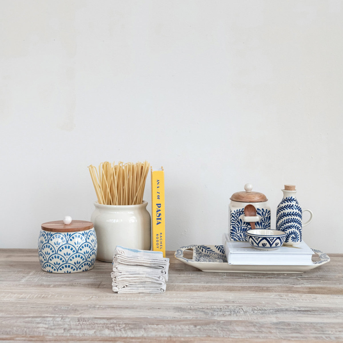The HAND-PAINTED STONEWARE CANISTER WITH LID AND SPOON by CREATIVE COOP features blue floral designs, an acacia wood lid, and a wooden spoon in a side holder—perfect as farmhouse kitchen decor. Sits on a white background.