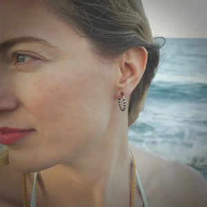 The ALICE RISE LITTLE COLORLOOP EARRINGS IN SLATE BLUE by ALICE RISE, featuring gold hoops with alternating white and teal Japanese glass beads, are displayed on a textured light blue surface, highlighting their handmade elegance.